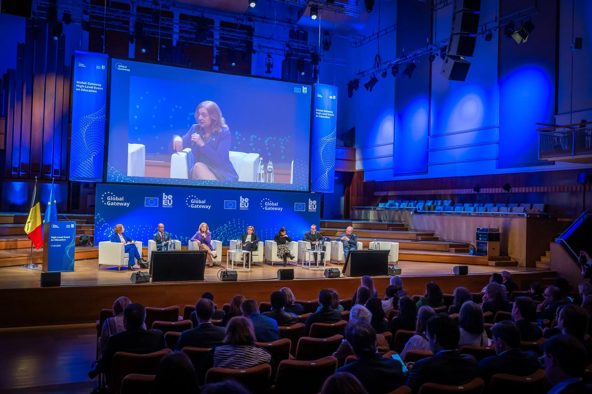 A conference with speakers on stage at a ‘Global Gateway’ event, featuring a giant screen showing a female speaker, with EU and Belgian flags on display.