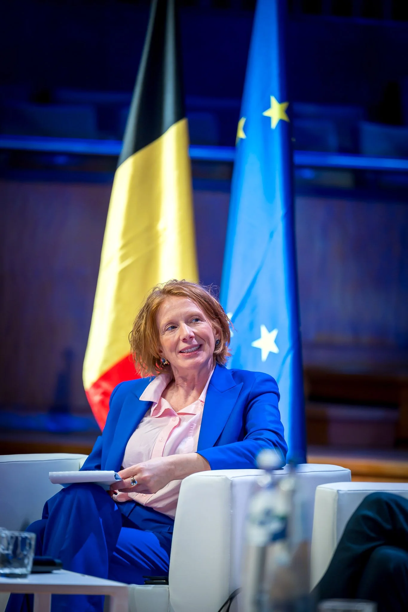 Une femme en costume bleu assise dans une salle avec des drapeaux de Belgique et de l'Union européenne en arrière-plan.