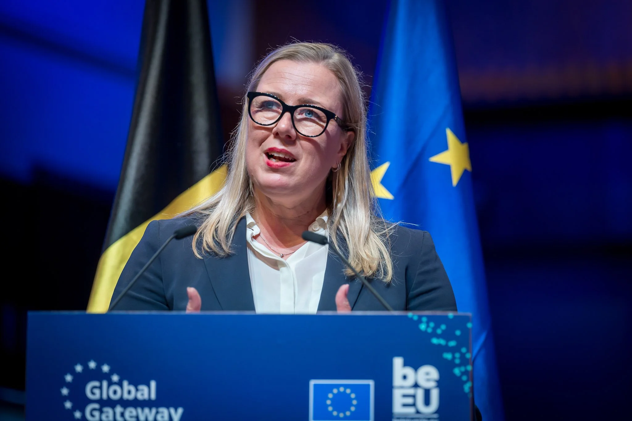 A blonde woman wearing glasses is speaking at a lectern bearing the words ‘Global Gateway’ and ‘be EU’, with the flags of Belgium and the European Union in the background.