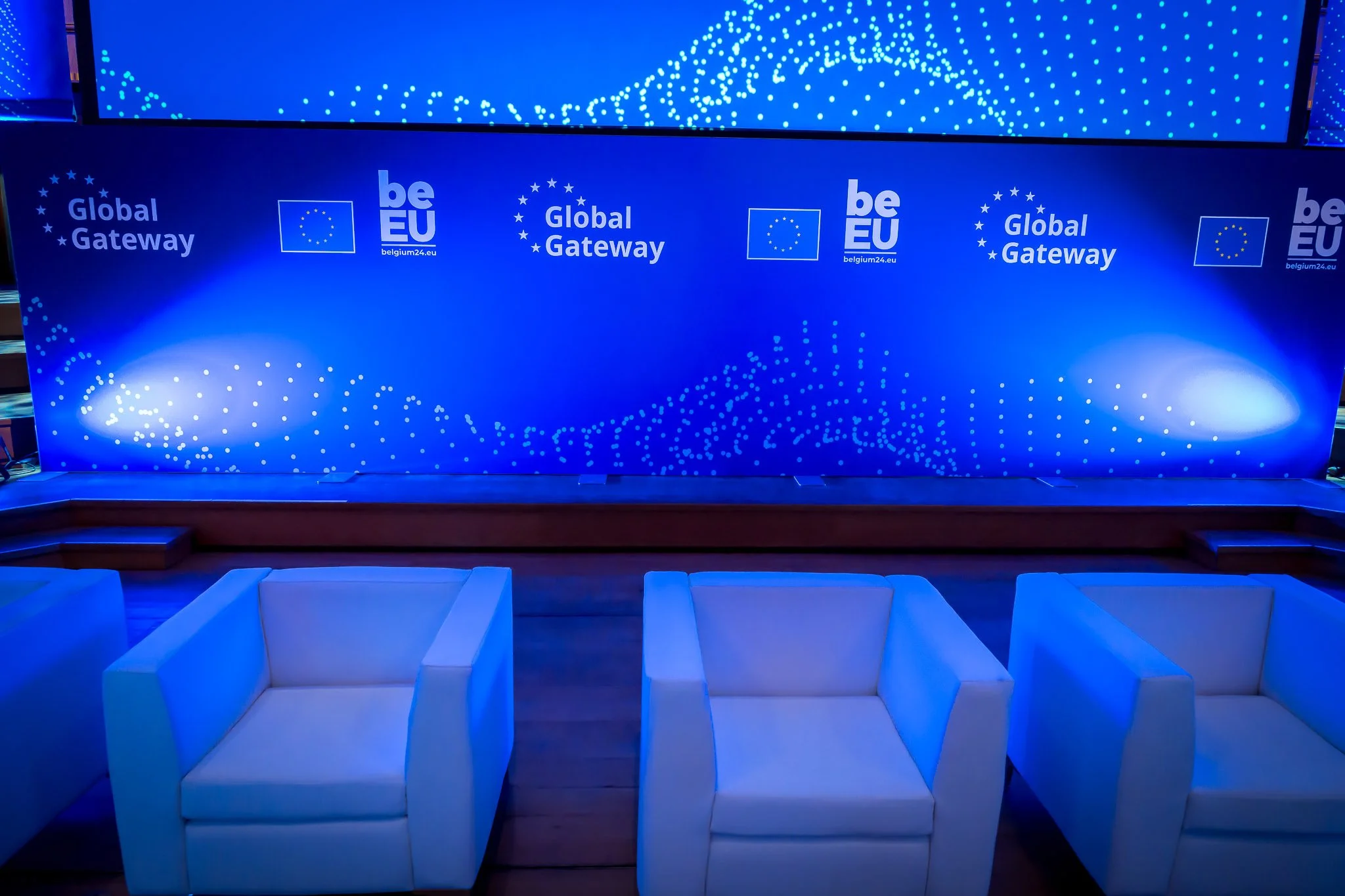 A scene featuring three white armchairs against a blue backdrop bearing the ‘be EU’ and ‘Global Gateway’ logos.