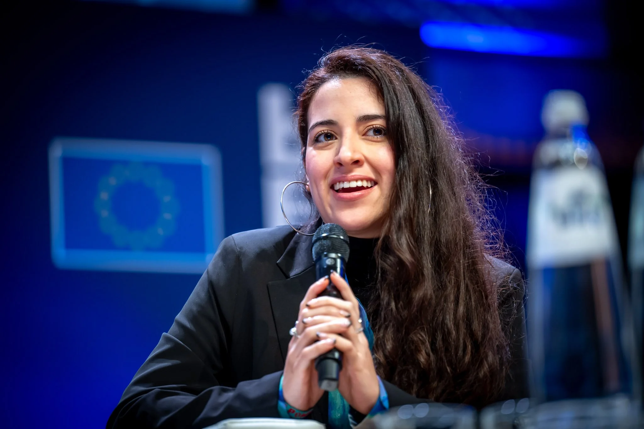 Jeune femme à la conférence, souriante, parlant dans un microphone, fond bleu avec logo de l'Union européenne.