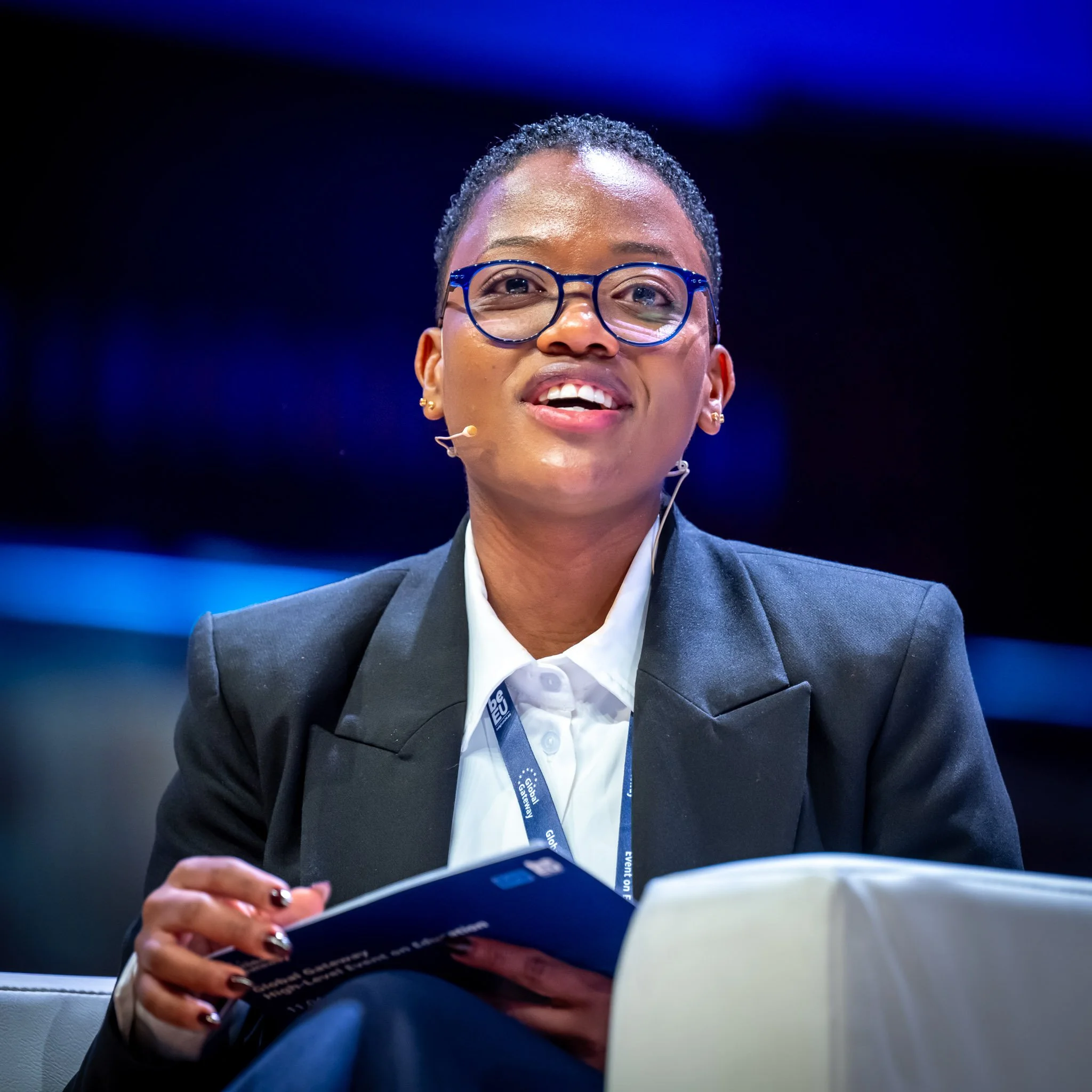 Femme souriante, portant des lunettes et une veste noire, tenant un guide ou un rapport lors d'un événement.