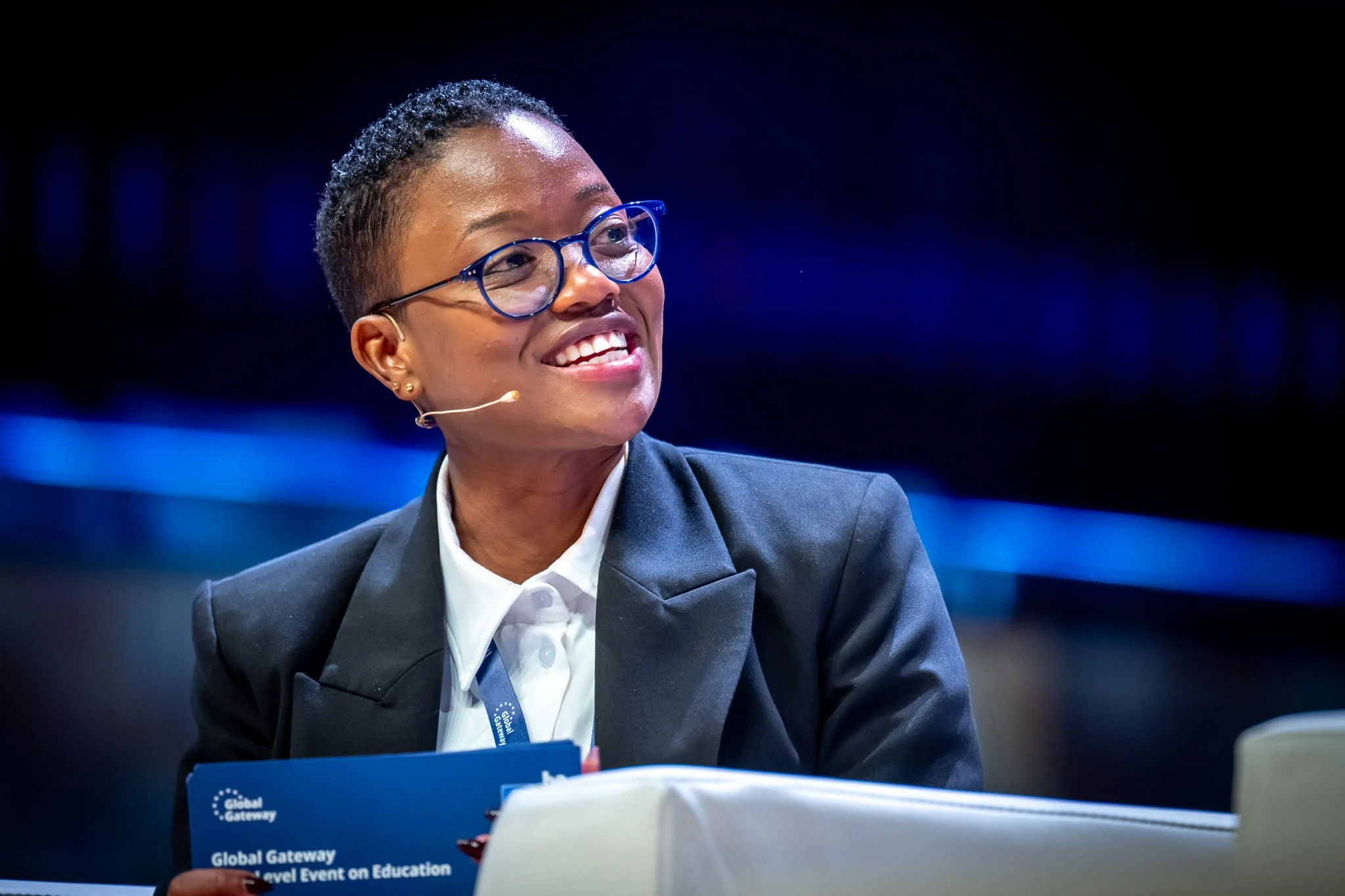 Une femme souriante, portant des lunettes bleues et un costume noir, assise lors d'une conférence ou d'un événement éducatif, tenant un livret bleu.