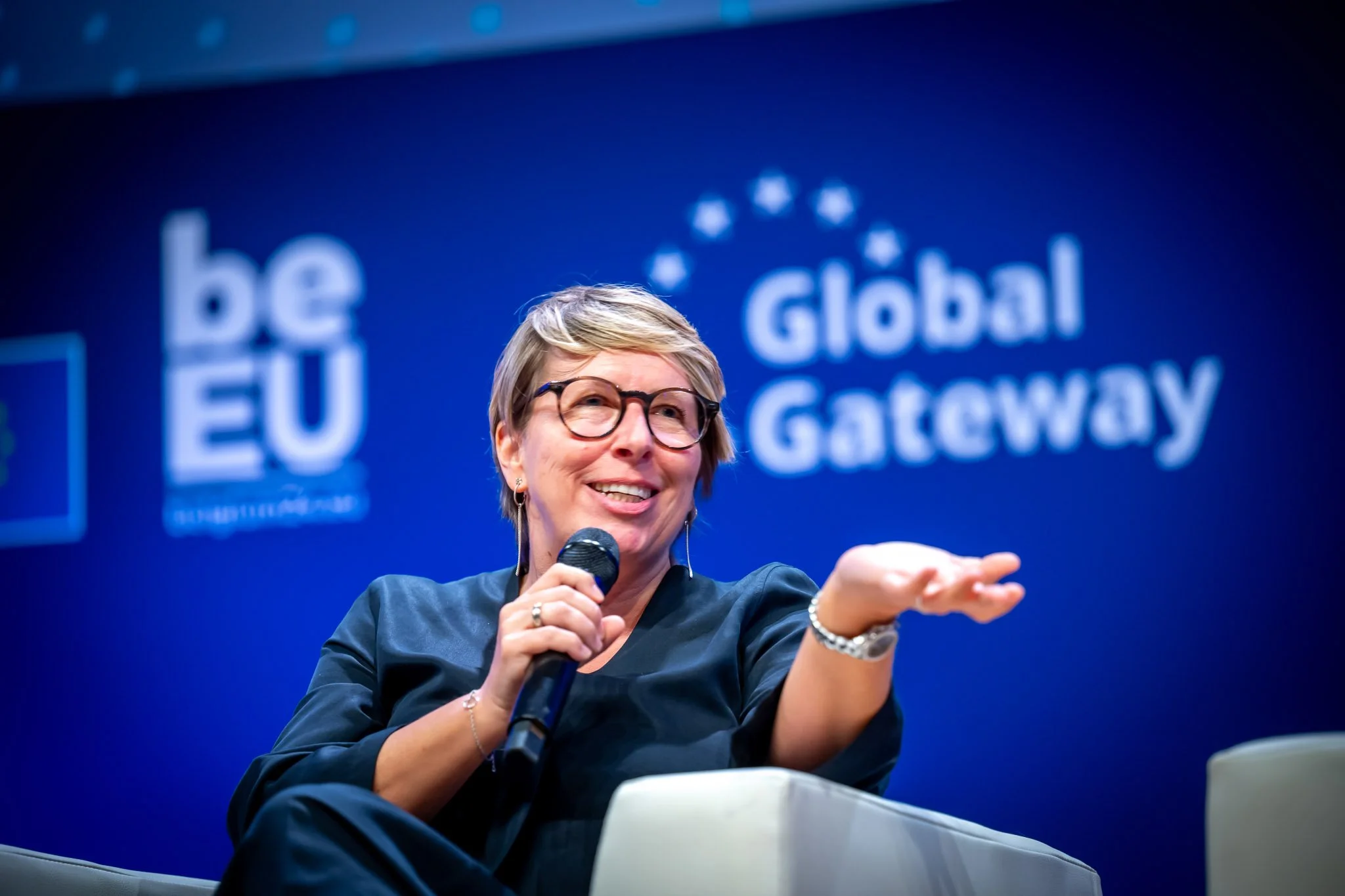 Photo professionnelle d'une ministre belge lors d'un événement international. La ministre tenant un micro avec une toile de fond bleue indiquant "Global Gateway" et "be EU". Elle semble en train de parler lors d'un événement.