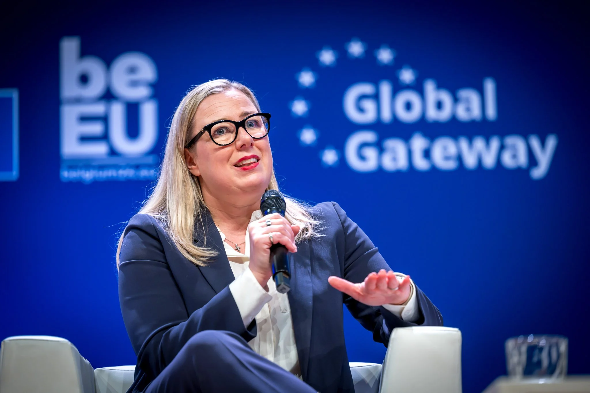 Une femme en costume, tenant un micro, parle lors d'un événement avec les mots "be EU" et "Global Gateway" en arrière-plan sur un écran bleu.