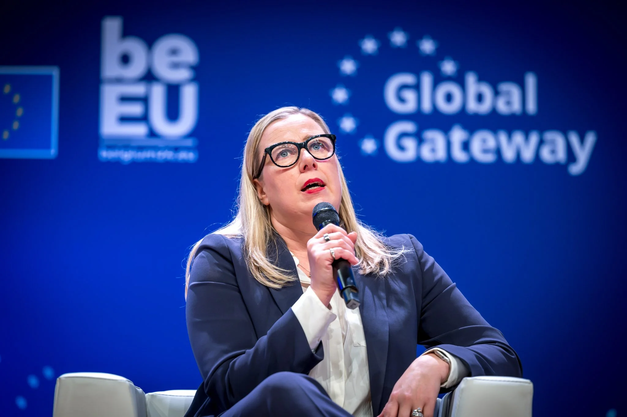 Une femme en costume bleu tenant un micro, assise devant un fond bleu avec les mots "Global Gateway" et "be EU" inscrits, lors d'un événement ou une conférence.