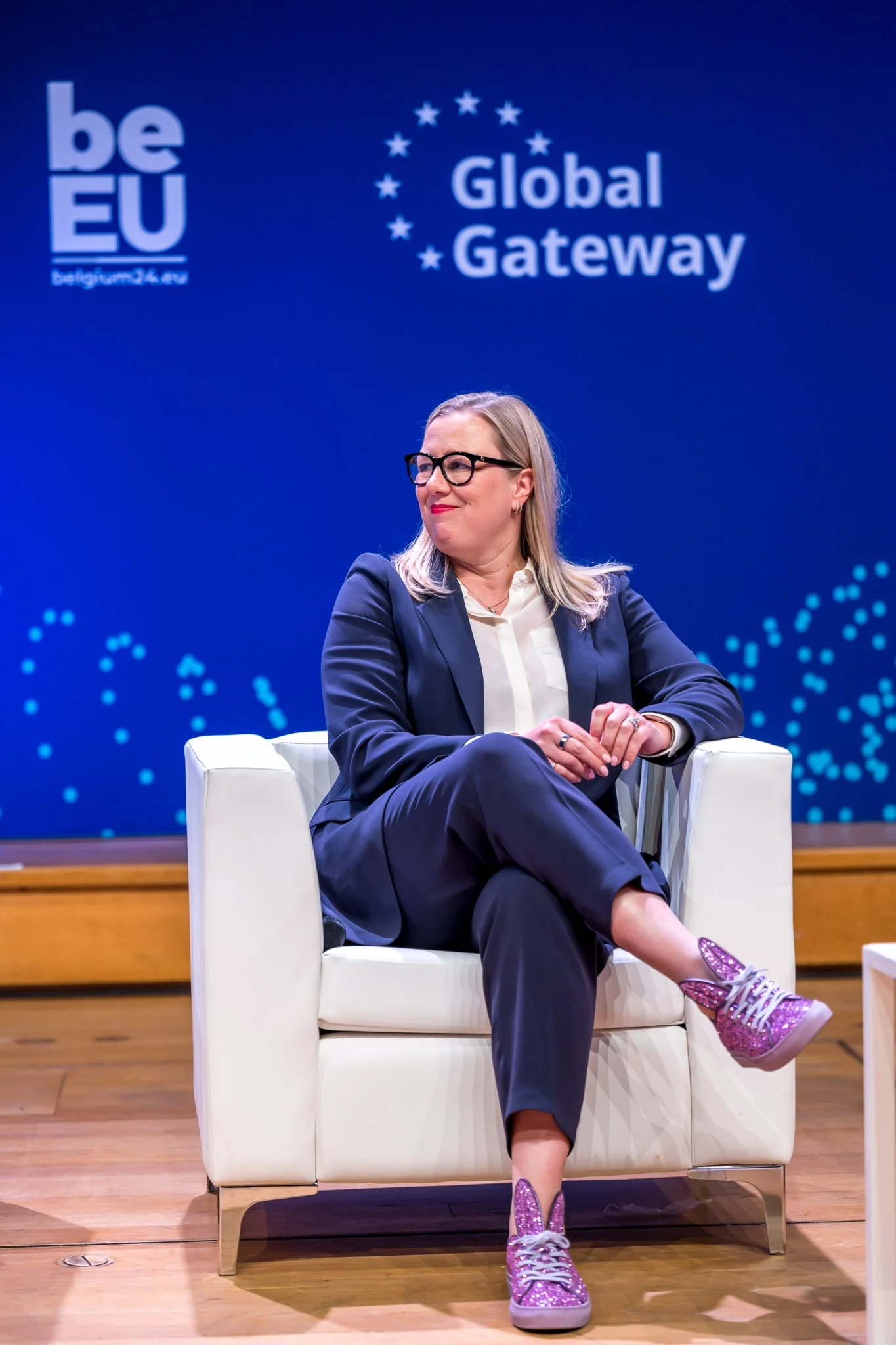 Une personne assise sur un fauteuil blanc, portant un costume bleu foncé et des baskets brillantes roses. En arrière-plan, on voit les logos 'be EU' et 'Global Gateway' sur un fond bleu.