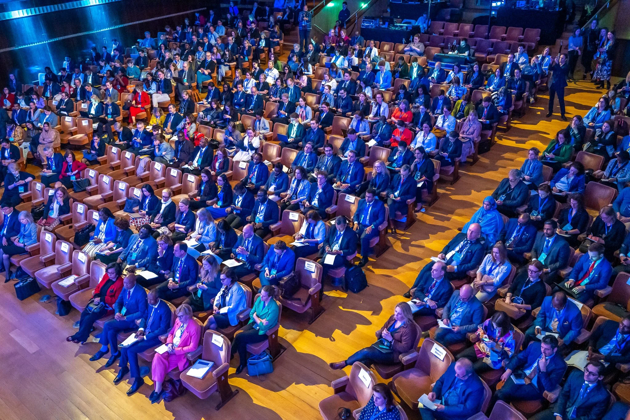 Photo d'une conférence. Vue aérienne d'une salle de conférence remplie de personnes assises, portant des vêtements formels, participant à un événement professionnel.