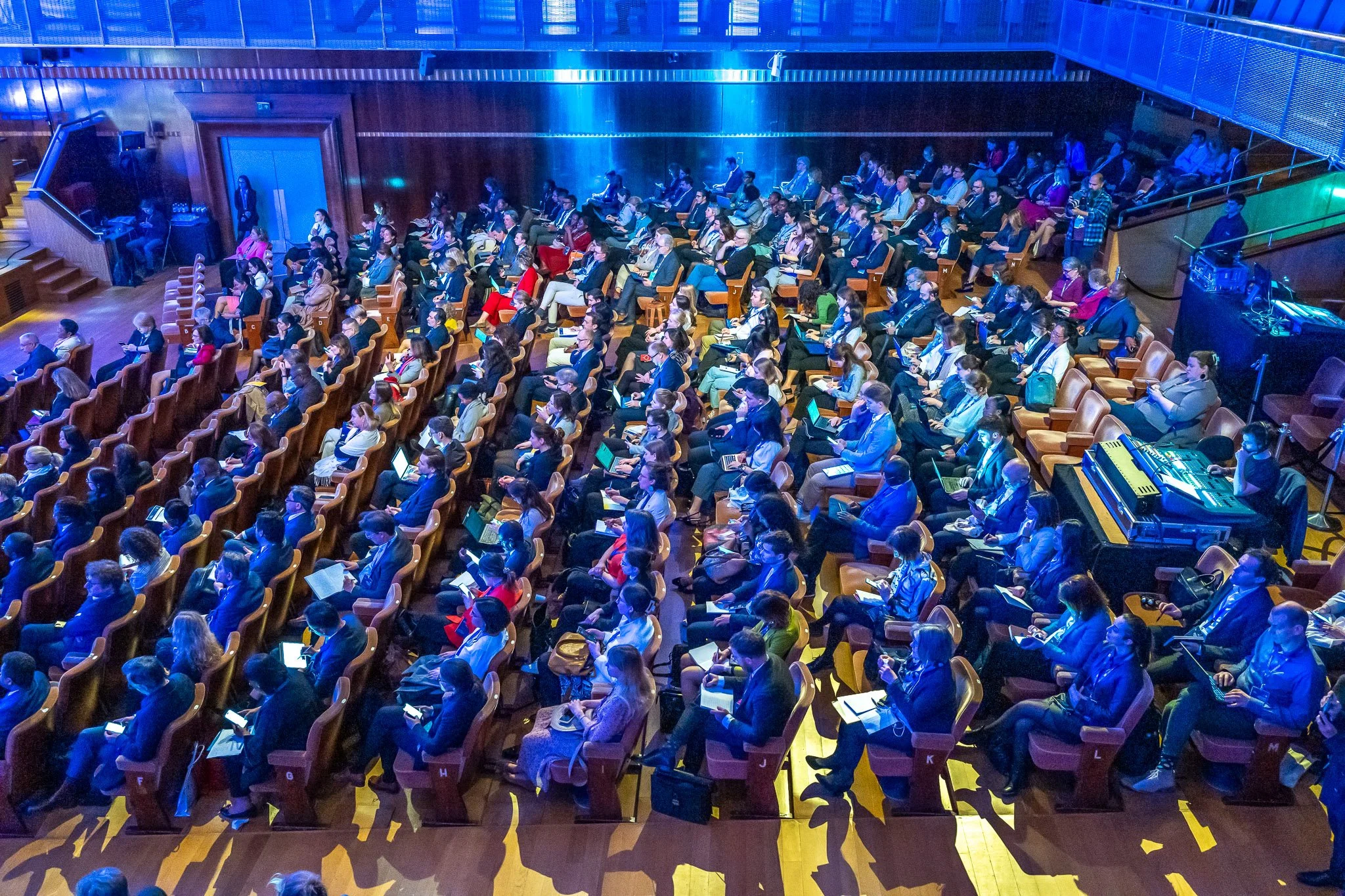 Une salle de conférence ou auditorium remplie de personnes assises dans des rangées de sièges en bois, utilisant des ordinateurs portables ou des tablettes, avec un éclairage tamisé et un système de sonorisation visible.