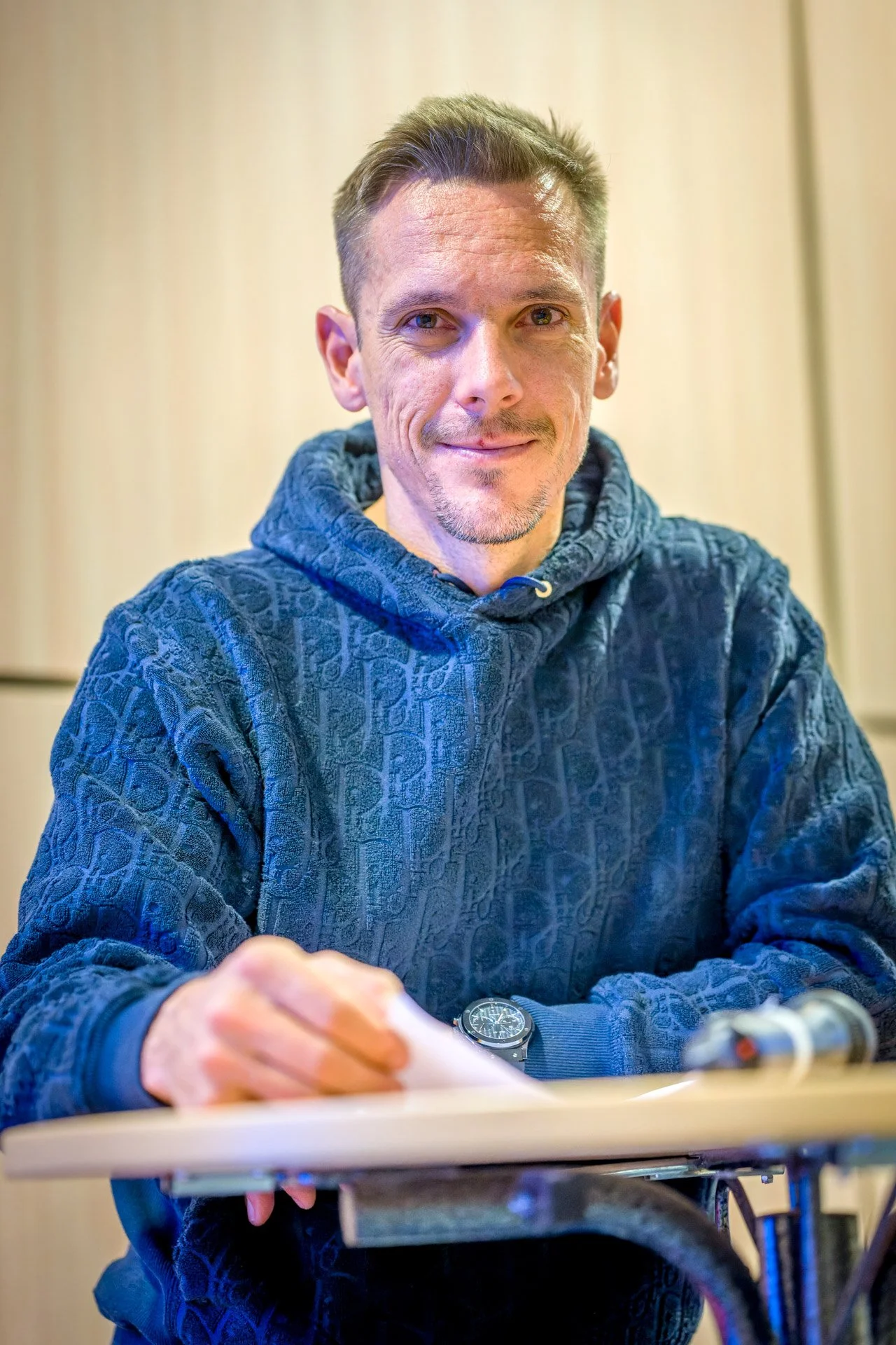 Un homme assis porte un pull bleu, tenant un papier et regardant la caméra, avec un fond flou beige.