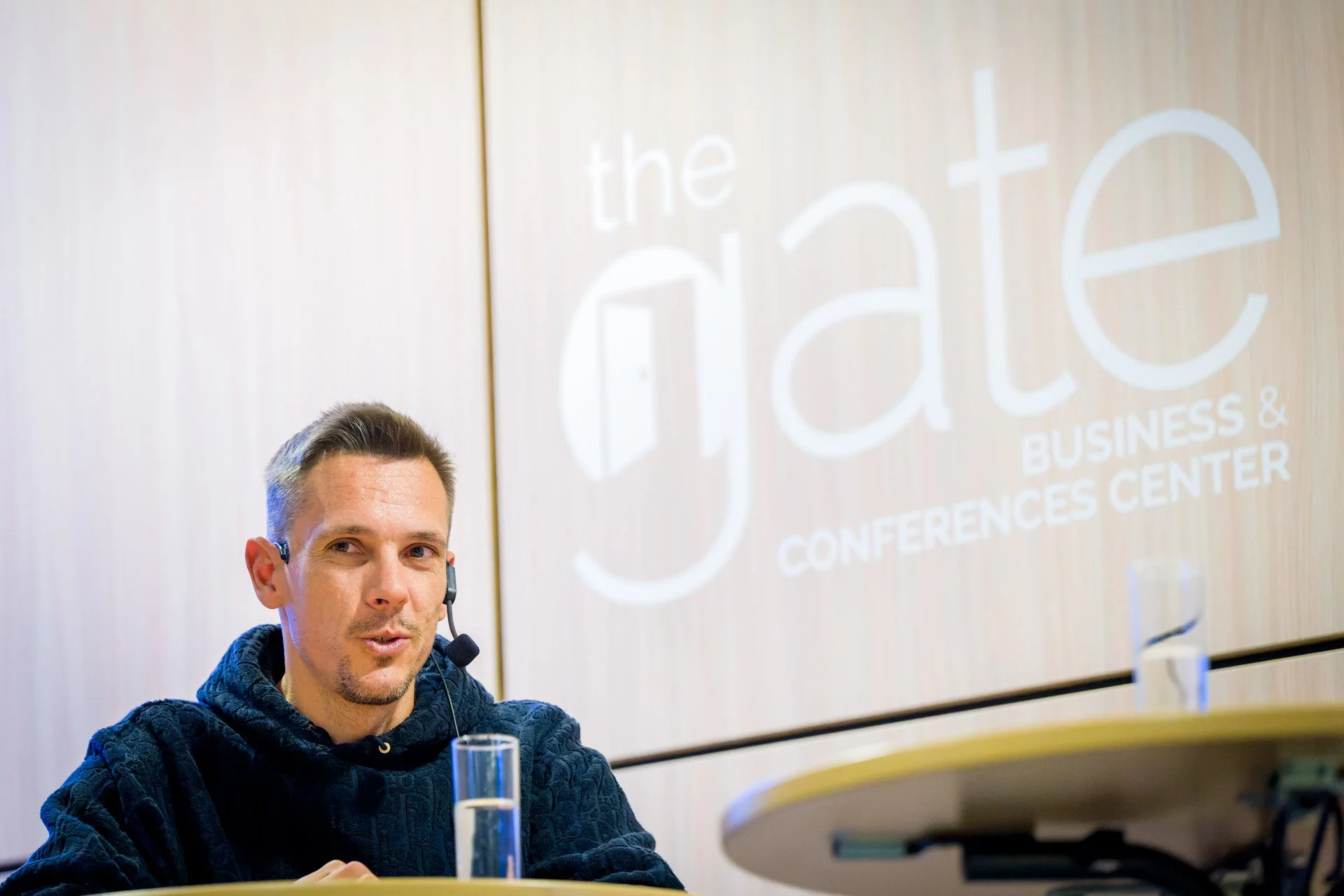 Un homme portant un casque micro parle devant une table avec un verre d'eau. En arrière-plan, le logo de "the gate Business & Conferences Center" est visible sur le mur.