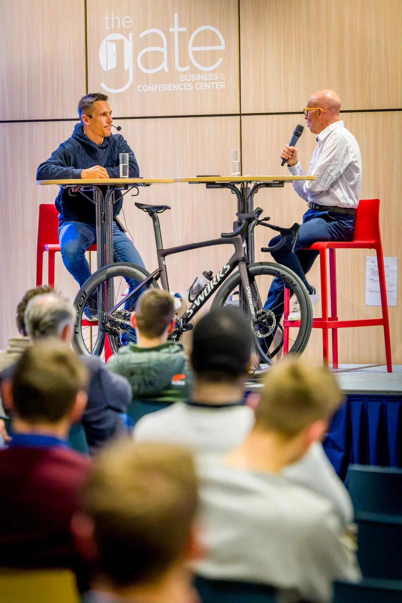 Deux hommes assis sur scène lors d'une conférence, discutant autour d'un vélo de course, avec un public en arrière-plan, au "Gate Business & Conferences Center".