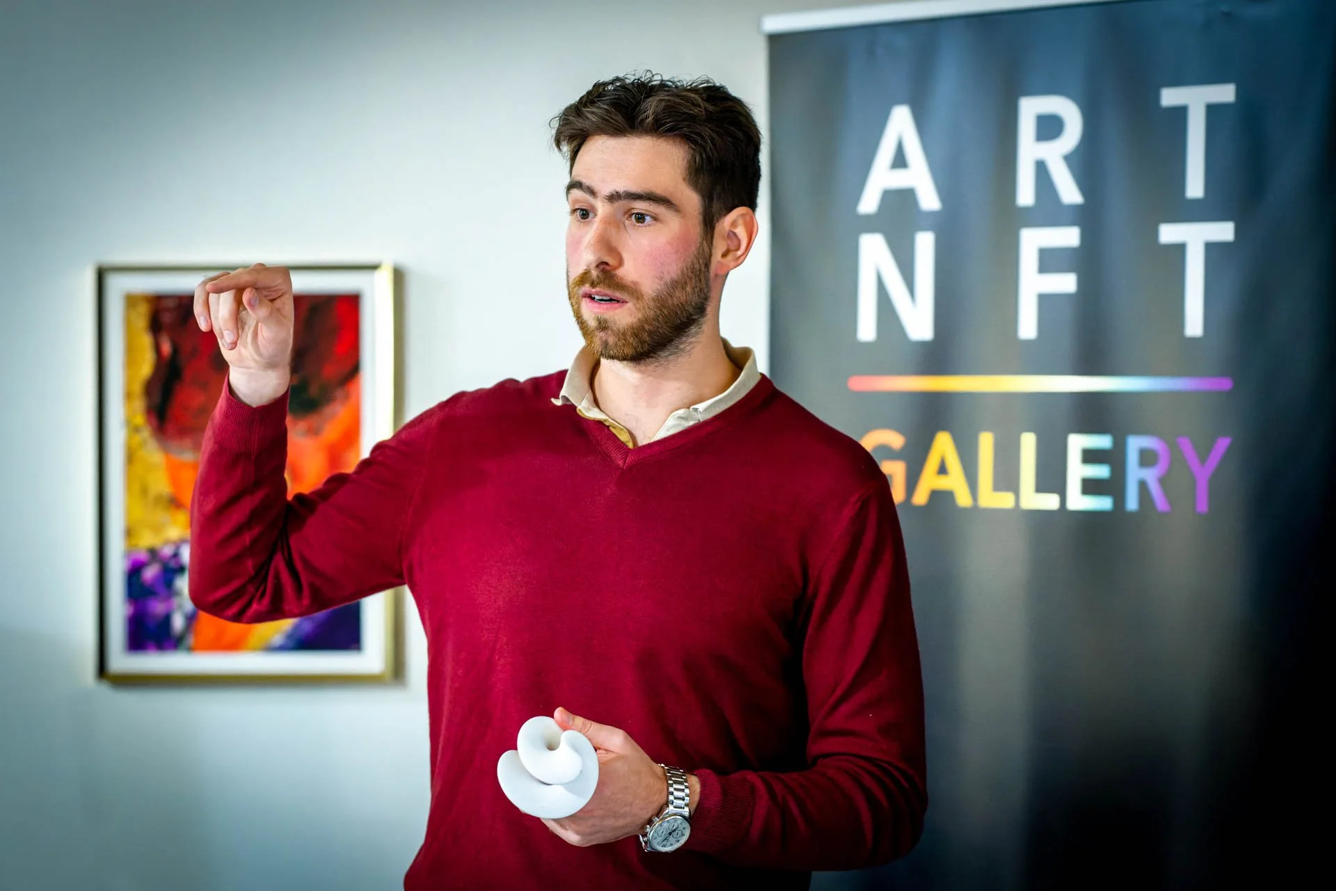 Un homme en pull rouge donne une présentation, tenant un objet blanc, devant un panneau "ART NFT GALLERY". En arrière-plan, on voit un tableau coloré."