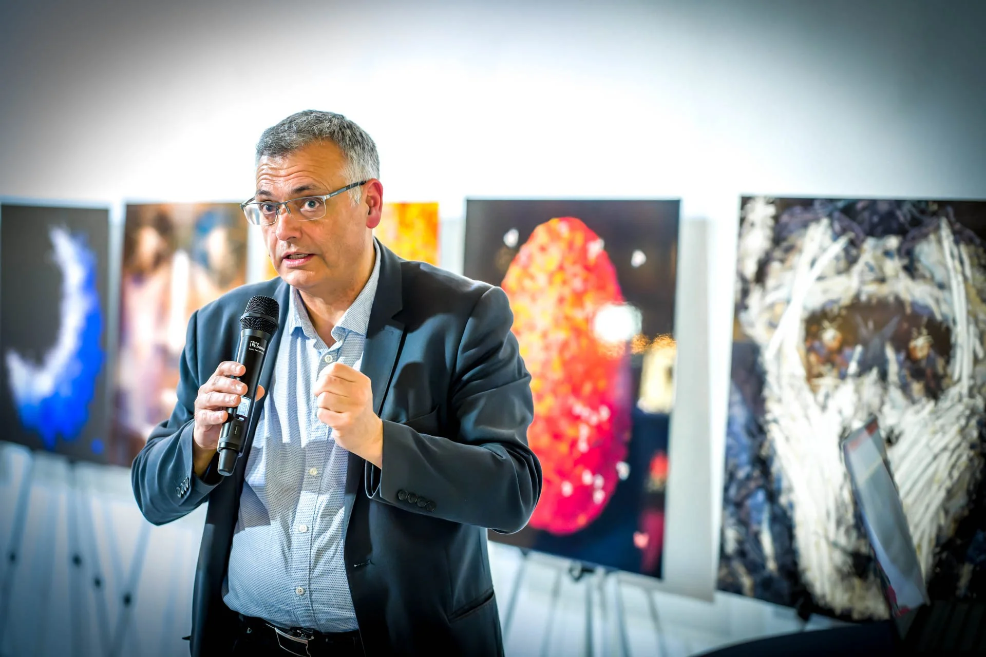 Un homme portant un costume noir parle lors d'une présentation ou d'une conférence dans une galerie d'art, avec des images artistiques en arrière-plan.