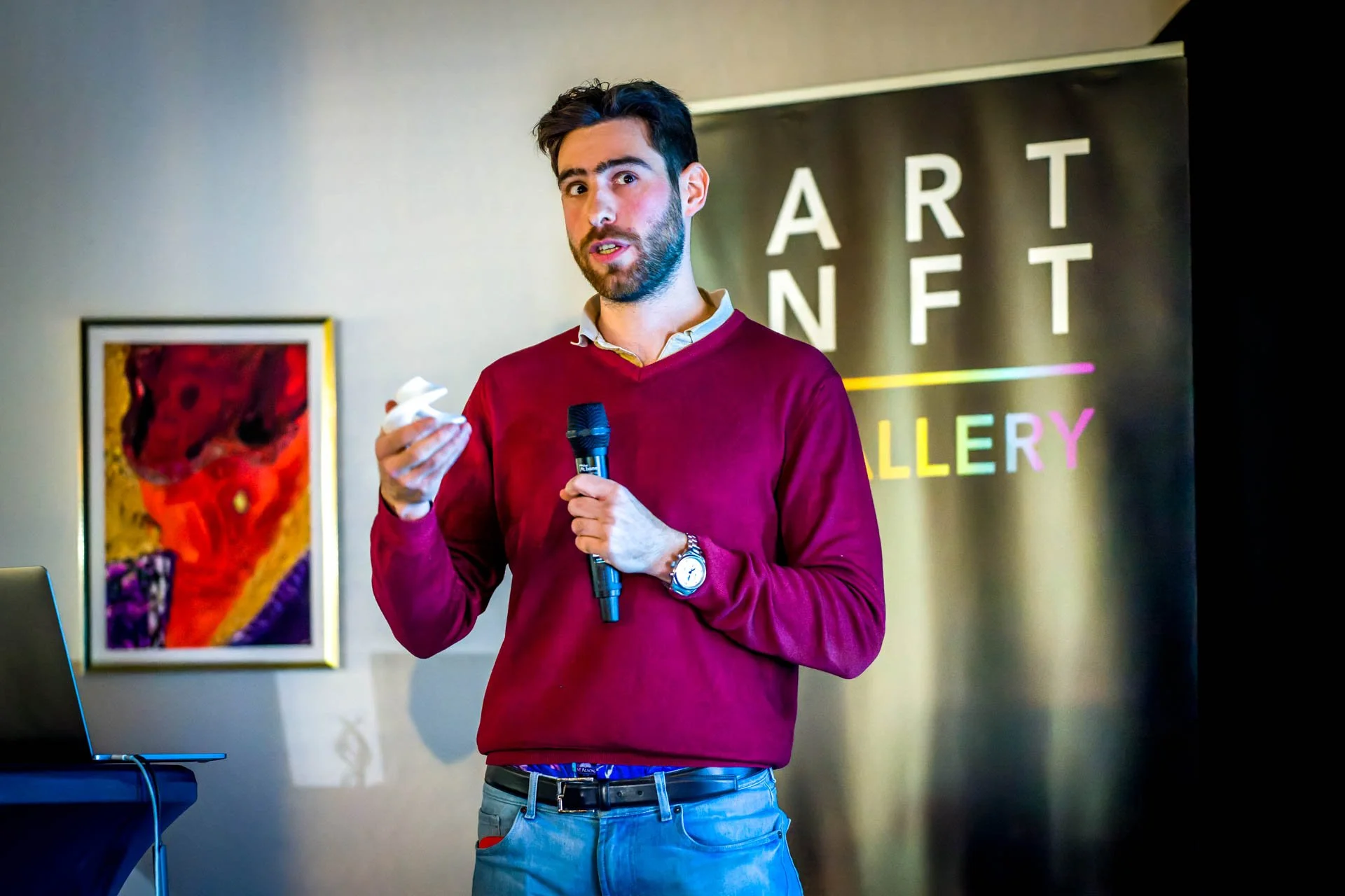 Un homme en pull rouge tient un micro et donne une présentation devant une bannière marquée "ART NFT GALLERY". Une peinture colorée est accrochée au mur en arrière-plan.