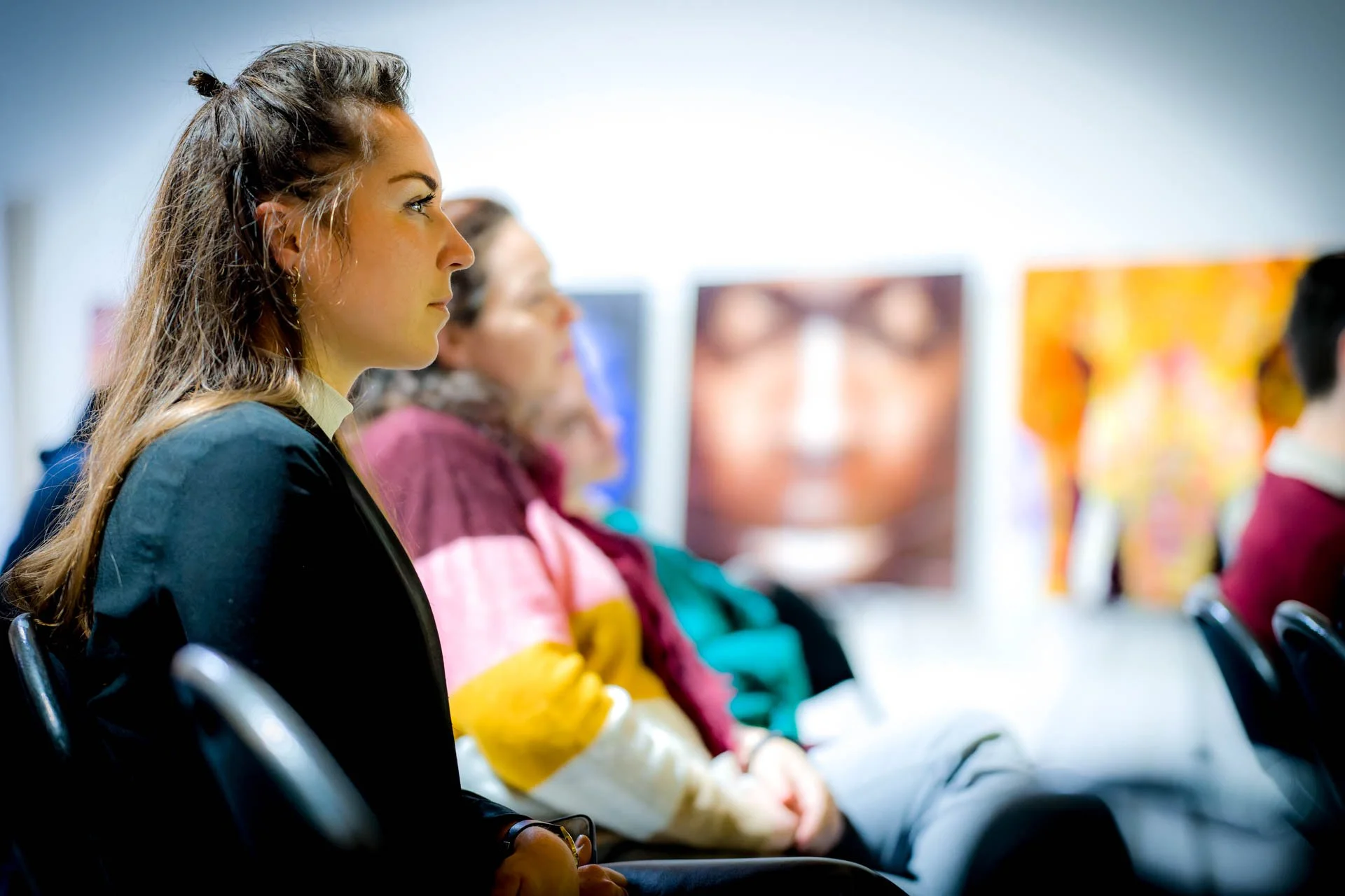 Personnes assises attentivement dans une galerie d'art, regardant vers l'avant. Œuvres floues en arrière-plan.
