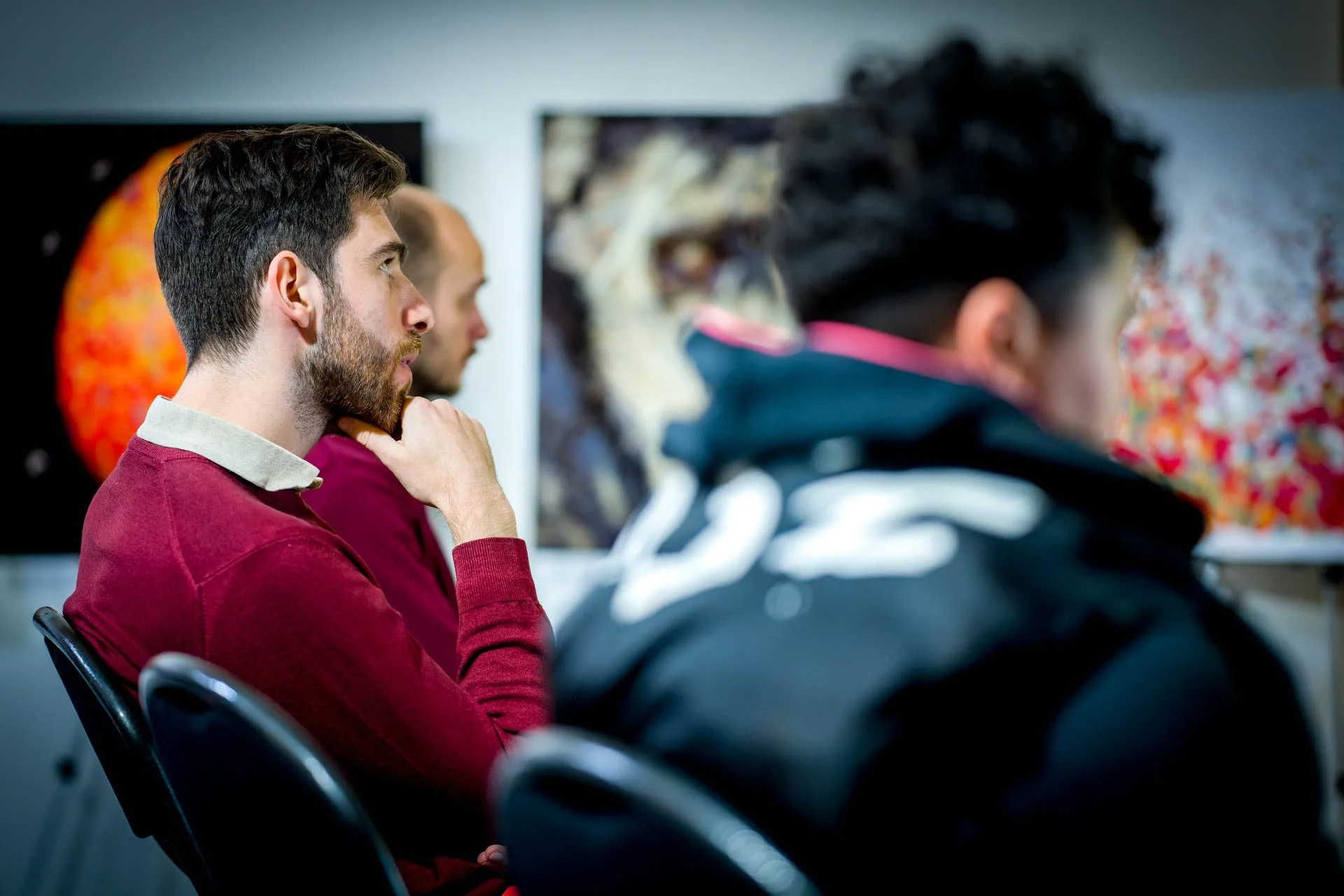 Des hommes assis écoutant attentivement dans une salle, avec des tableaux colorés en arrière-plan.
