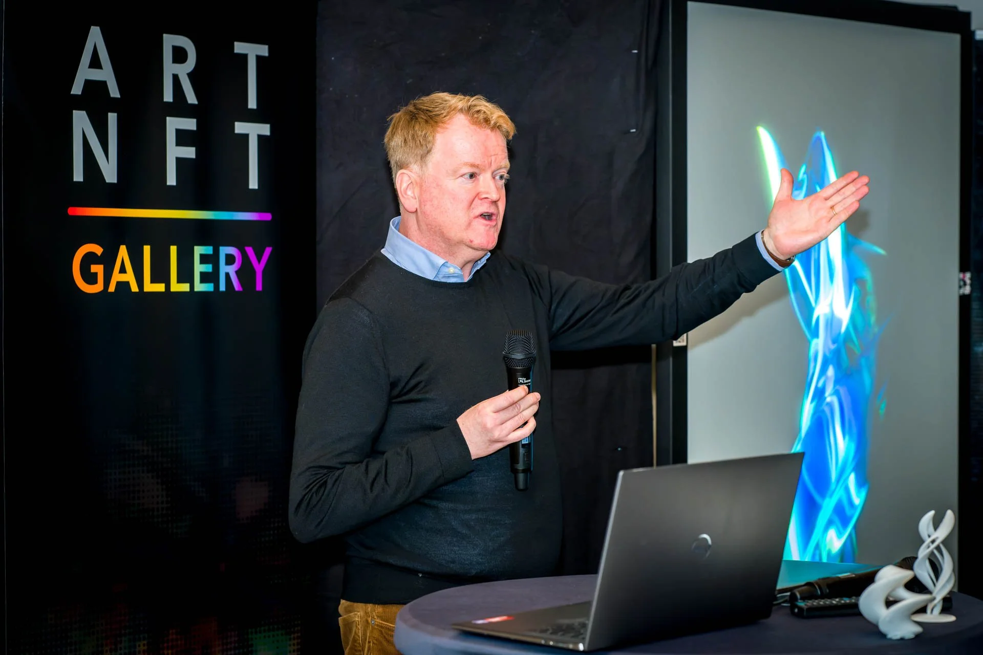 Un homme tenant un micro présente devant un écran montrant une image numérique abstraite, avec une pancarte "ART NFT GALLERY" en arrière-plan.