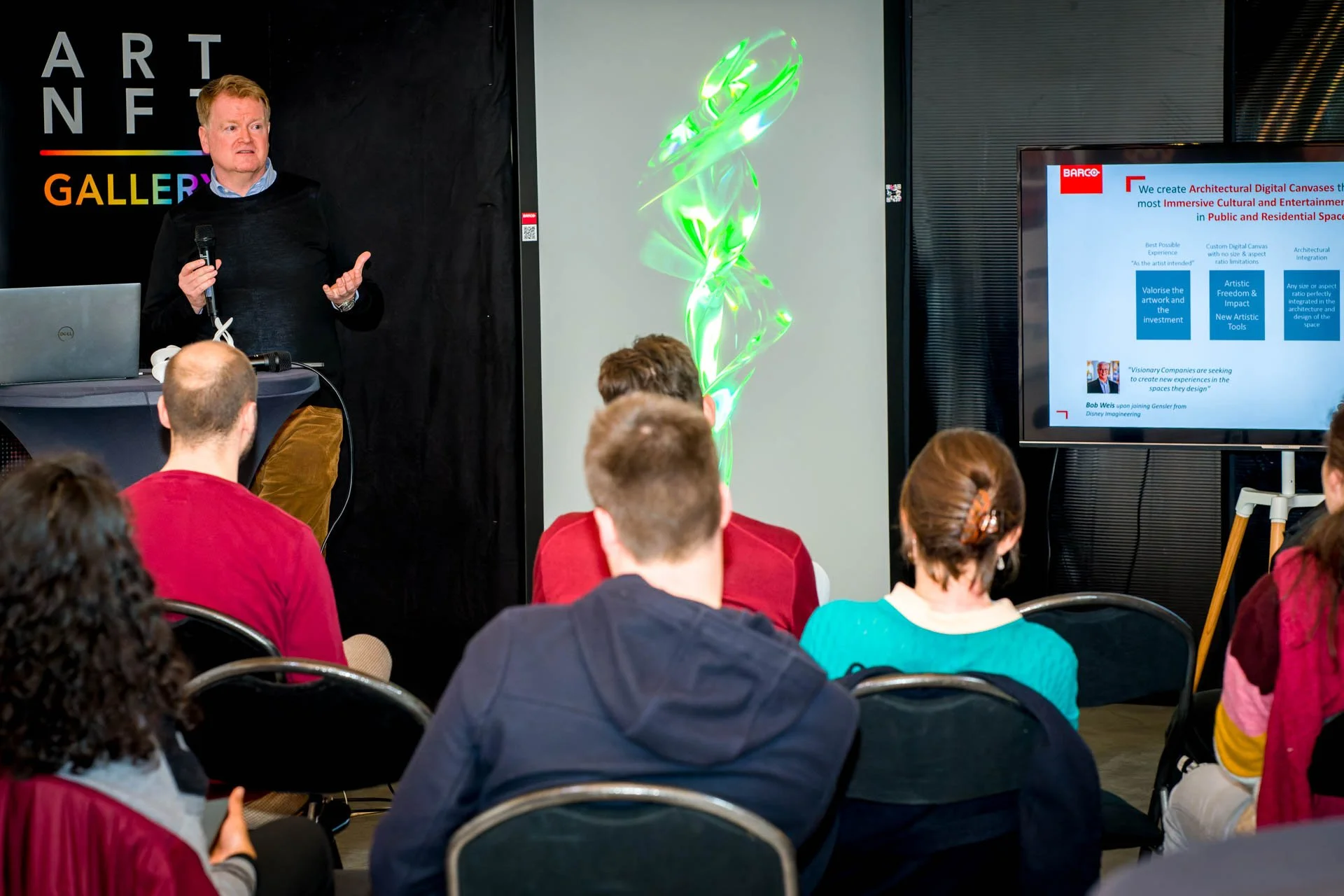 Un homme fait une présentation devant un public assis, avec un écran affichant une diapositive et un objet lumineux vert en arrière-plan. Une enseigne "ART NFT GALLERY" est visible.
