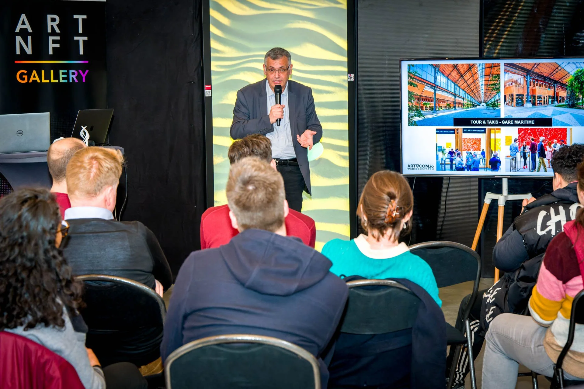 Un homme fait une présentation devant un public assis. Il tient un microphone. Il y a un écran affichant des images d'un bâtiment dénommé "Tour & Taxis - Gare Maritime". Un panneau indique "ART NFT GALLERY" à gauche. L'événement semble lié aux NFT ou