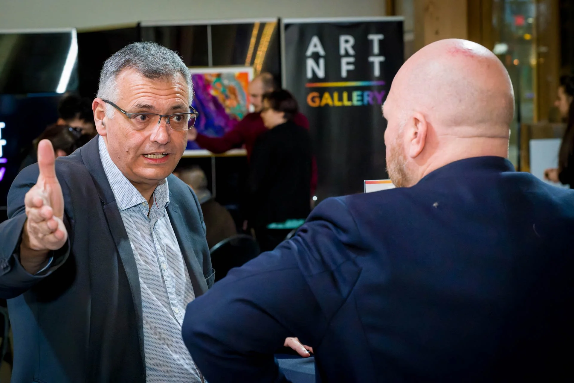Deux hommes discutant devant une exposition d'art NFT, un homme a l'air sérieux avec des lunettes et l'autre de dos. Arrière-plan avec des affiches et des œuvres d'art colorées.