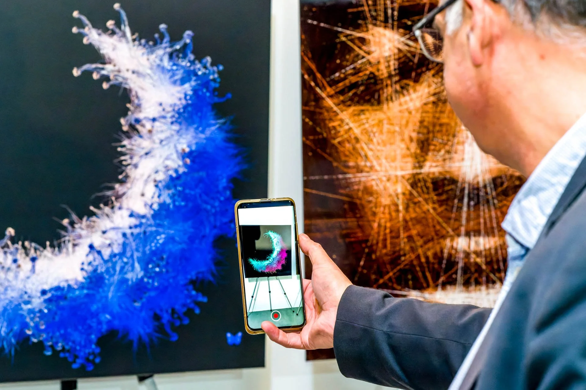 Un homme photographie une œuvre d'art numérique avec son téléphone, l'œuvre représente une structure colorée en forme de C en train de se déployer.