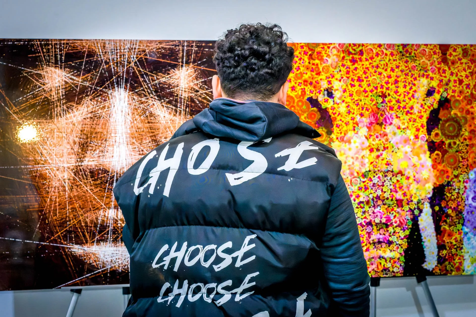 Photo d'un événement organisé dans une galerie d'art à Bruxelles. Un homme de dos portant une veste avec l'inscription "CHOOSE" devant une œuvre d'art abstraite colorée sur un mur.