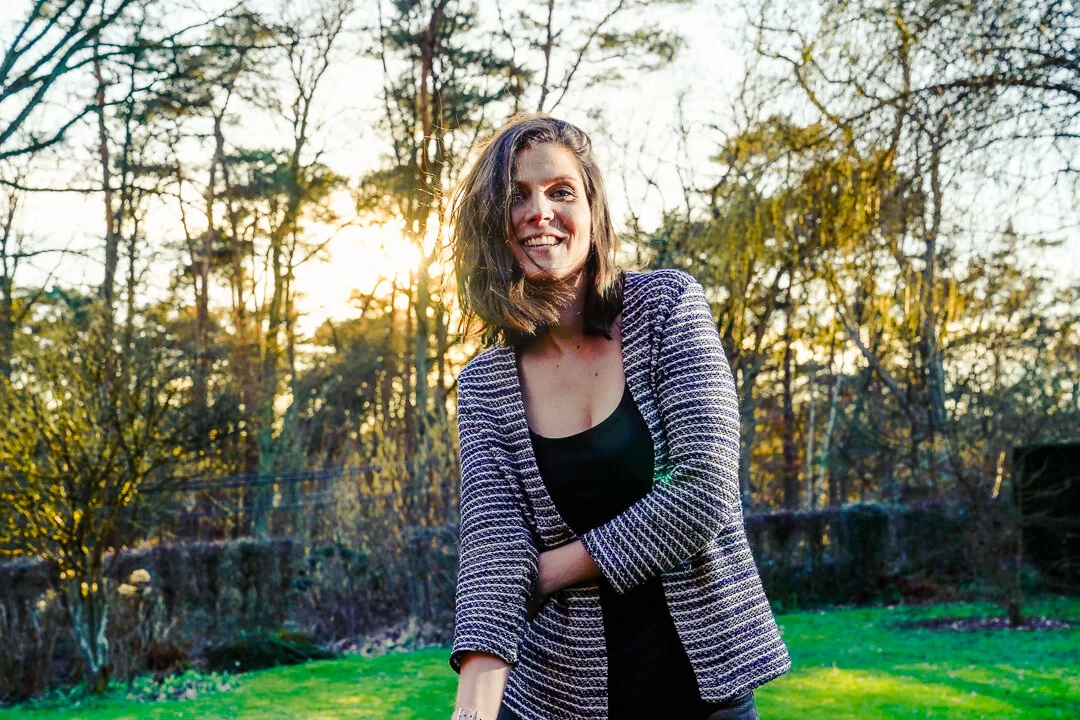 Une femme sourit dans un jardin, devant des arbres éclairés par le soleil couchant.